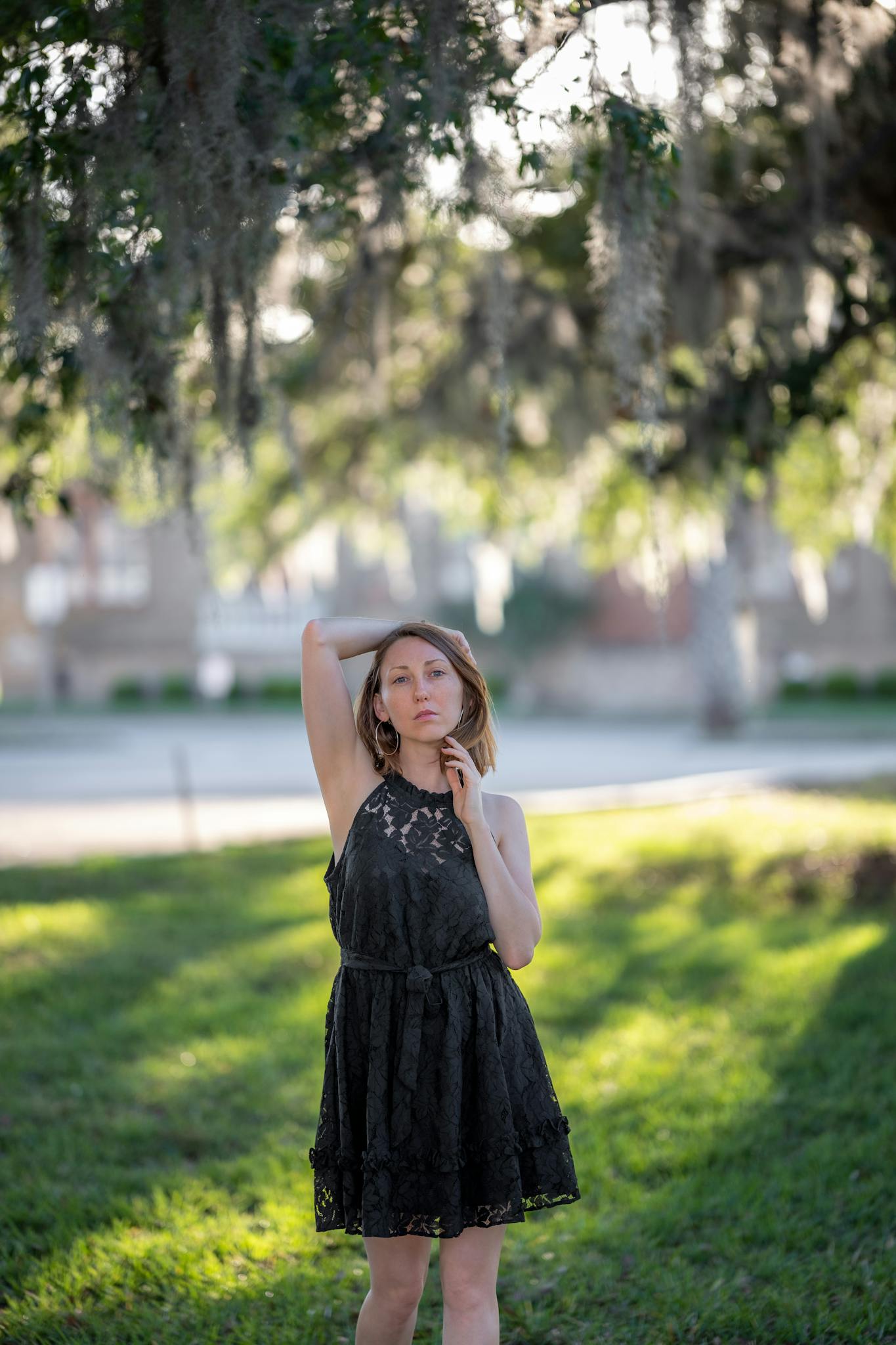A young woman elegantly poses outdoors in a stylish black dress under hanging foliage.